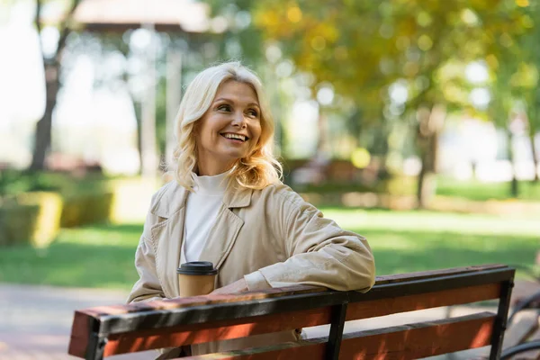 Glückliche Frau mittleren Alters hält einen Kaffee to go in der Hand, während sie auf einer Bank im verschwommenen Park sitzt — Stockfoto