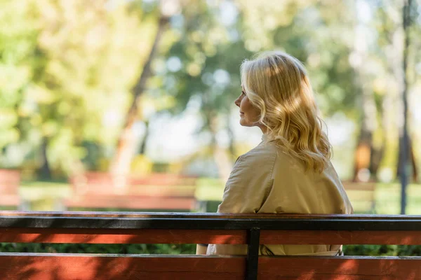 Vista lateral de la mujer madura en gabardina sentado en el banco en el parque - foto de stock