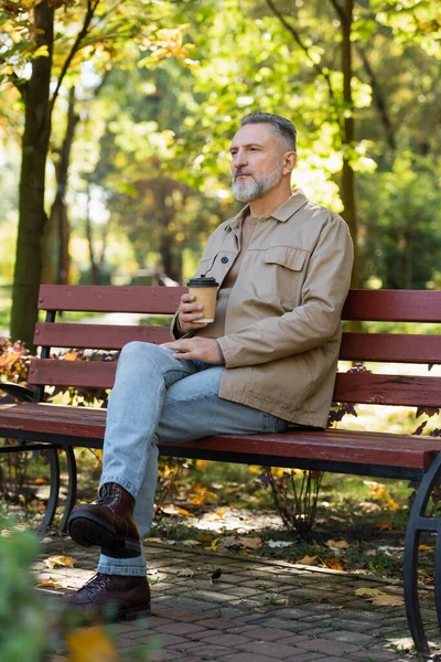 Зрелый мужчина держит кофе, чтобы пойти, сидя на скамейке в парке — стоковое фото