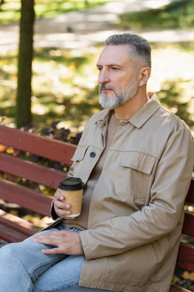 Зрелый мужчина в весеннем наряде держит кофе, чтобы пойти, сидя на скамейке в парке — стоковое фото
