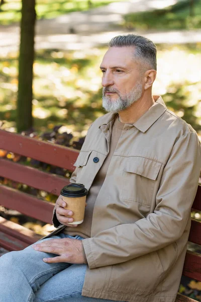 Mature man in spring outfit holding coffee to go while sitting on bench in park — Stock Photo