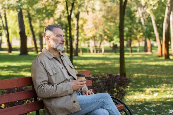 Hombre de mediana edad sosteniendo taza de papel mientras pasa tiempo en el banco en el parque de primavera - foto de stock