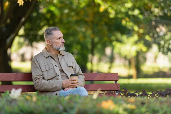 Мужчина средних лет держит выпивку на вынос и смотрит в сторону, сидя на скамейке в парке — стоковое фото