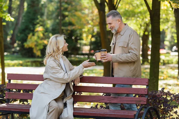 Вид сбоку улыбающегося мужчины с бумажными чашками рядом с женой блондинкой, сидящей на скамейке в парке — стоковое фото