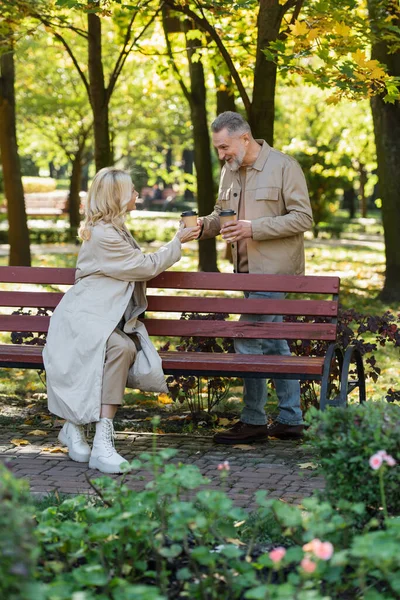 Sonriente hombre dando taza de papel con café a la esposa rubia en el banco en el parque - foto de stock