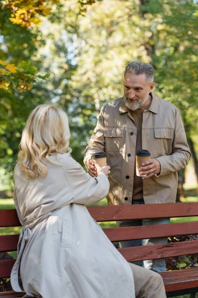 Mature husband giving coffee to go to blonde wife in trench coat in park — Stock Photo