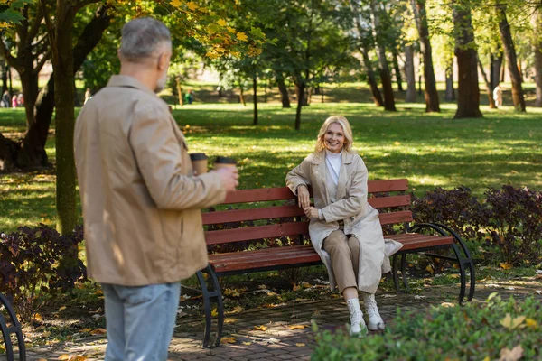 Smiling blonde woman looking at blurred husband with coffee to go in spring park — Stock Photo
