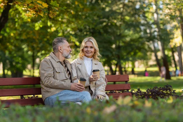 Femme insouciante tenant du café pour aller près du mari sur le banc dans le parc — Photo de stock