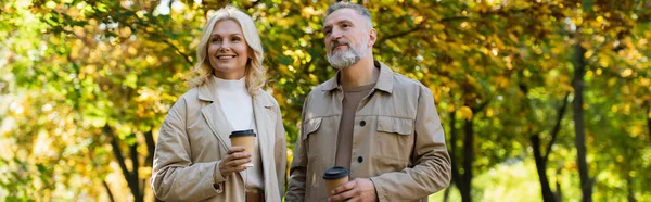 Безтурботний зріла пара тримає каву, щоб піти під час прогулянки в парку, банер — стокове фото