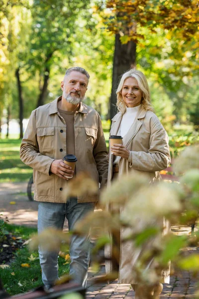 Усміхнена жінка в траншеї пальто тримає каву, щоб підійти до чоловіка в парку — стокове фото