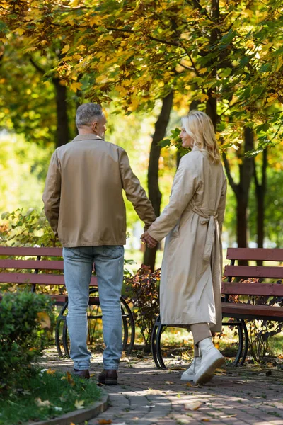Беззаботная женщина в плаще держит мужа за руку во время прогулки в парке — стоковое фото