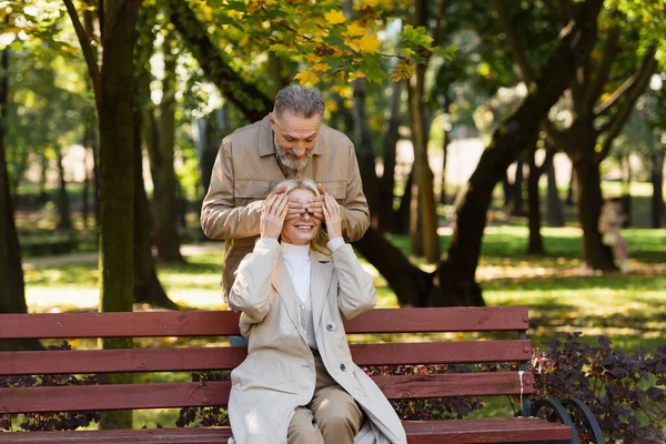 Улыбающийся мужчина, закрывающий глаза жене, сидящей на скамейке в парке — стоковое фото