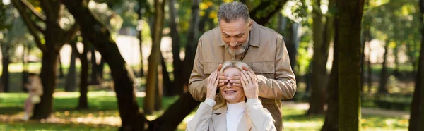 Reifer Mann bedeckt Augen von fröhlicher Frau im Park, Banner — Stockfoto