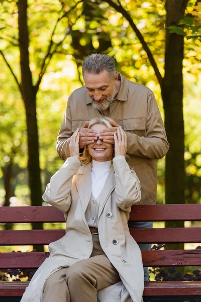 Positive mature man covering eyes of wife sitting on bench in park — Stock Photo