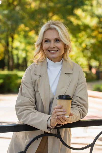 Retrato de mujer de mediana edad positiva sosteniendo café para ir en el parque de primavera - foto de stock