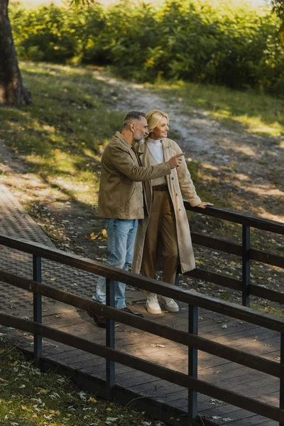 Smiling mature man pointing with finger near blonde wife while standing on bridge in park — Stock Photo