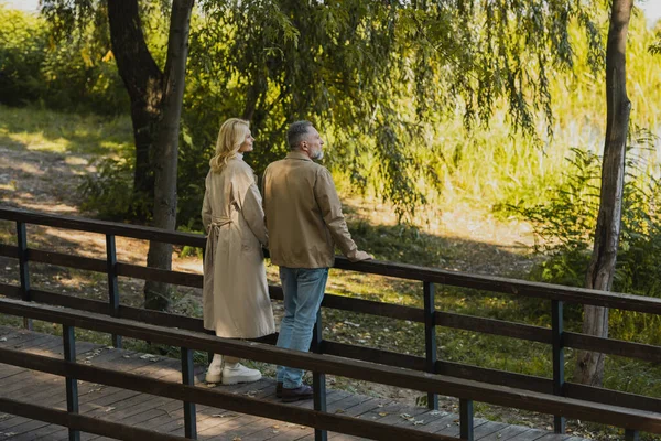 Vue latérale du couple d'âge moyen regardant loin tout en se tenant sur le pont dans le parc de printemps — Photo de stock