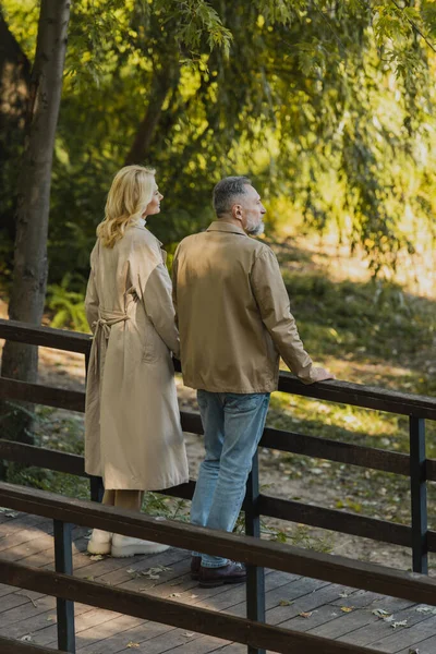 Vista lateral do casal de meia idade em roupas de primavera em pé na ponte no parque — Fotografia de Stock
