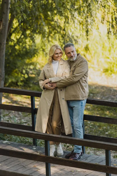 Reifer Mann umarmt unbeschwerte blonde Frau auf Brücke im Frühlingspark — Stockfoto