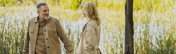 Sorridente uomo maturo guardando la moglie bionda in trench nel parco, banner — Foto stock