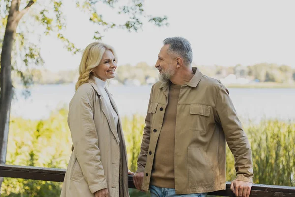 Sorrindo mulher loira em casaco de trincheira olhando para o marido maduro na ponte no parque — Fotografia de Stock