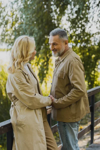 Lächelnder älterer Mann hält die Hand seiner blonden Frau, während er auf einer Brücke im Park steht — Stockfoto