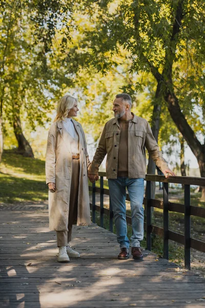 Seitenansicht eines Paares mittleren Alters, das auf einer Brücke im Park spricht und Händchen hält — Stockfoto