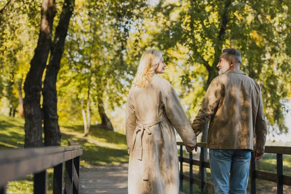 Вид на зрелую пару, держащуюся за руки во время прогулки по мосту в парке — стоковое фото