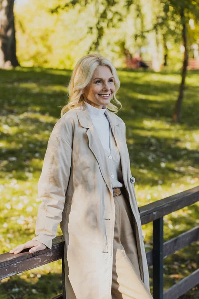 Cheerful blonde woman in trench coat standing on bridge in park — Stock Photo