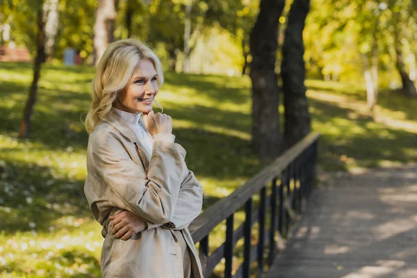 Беззаботная блондинка в плаще стоит на мосту в парке — стоковое фото