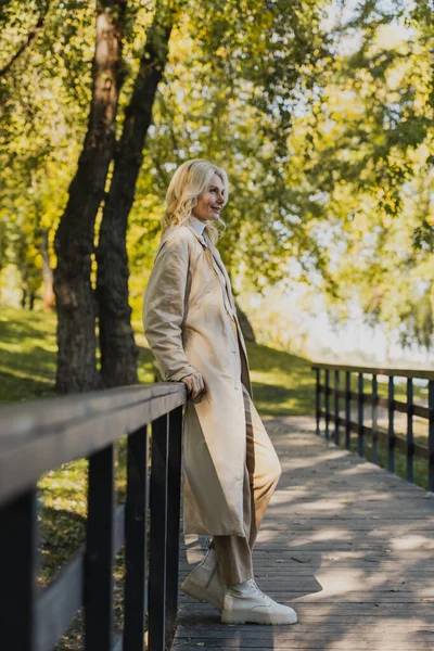 Mature blonde woman in trench coat smiling while standing on bridge in park — Stock Photo