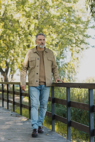 Пожилой человек в наружной одежде и джинсах, идущий по мосту в весеннем парке — стоковое фото