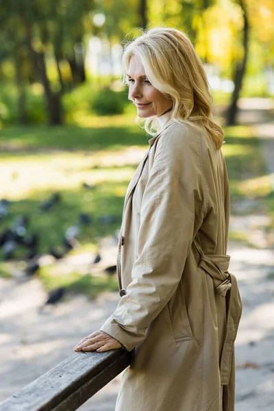 Overjoyed blonde woman in trench coat standing on bridge in spring park — Stock Photo