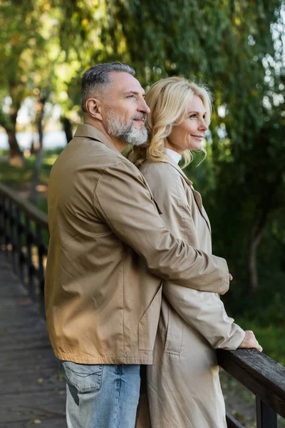 Smiling mature man hugging wife in trench coat while standing on bridge in park — Stock Photo