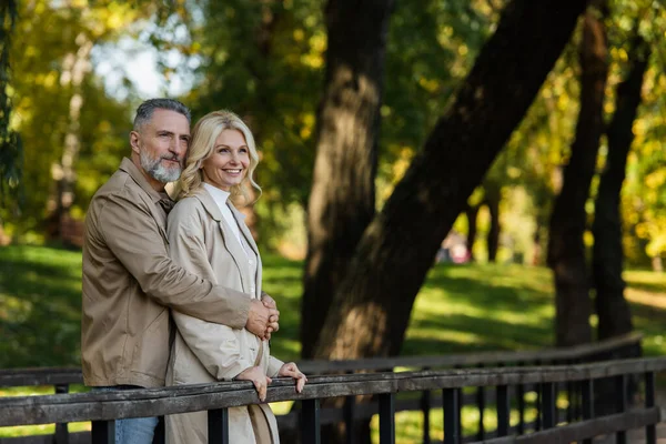 Mature man hugging smiling wife while standing on bridge in spring park — Stock Photo