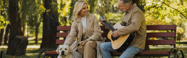 Мужчина играет на акустической гитаре возле беззаботной жены и лабрадора в парке, баннер — стоковое фото