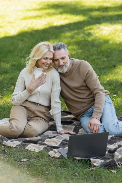 Encantador casal de meia-idade assistindo filme no laptop e sentado em cobertor no parque — Fotografia de Stock