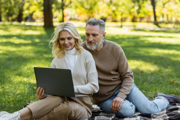 Despreocupado casal de meia idade assistindo filme no laptop e sentado em cobertor no parque — Fotografia de Stock