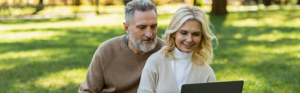Carefree middle aged couple watching movie on laptop in park during springtime, banner — Stock Photo