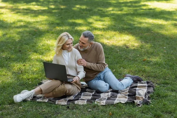 Despreocupado marido e mulher de meia-idade assistindo filme no laptop e sentado em cobertor no parque — Fotografia de Stock