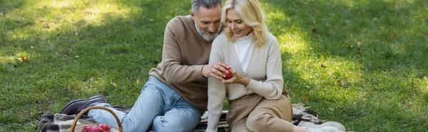 Щасливий чоловік і дружина тримають червоне яблуко і сидять на ковдрі під час пікніка в парку, банер — стокове фото