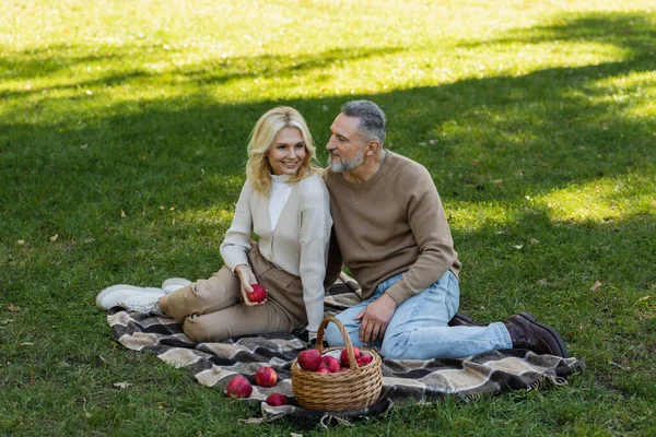 Довольная женщина средних лет, держащая спелое яблоко возле бородатого мужа во время пикника в парке — стоковое фото