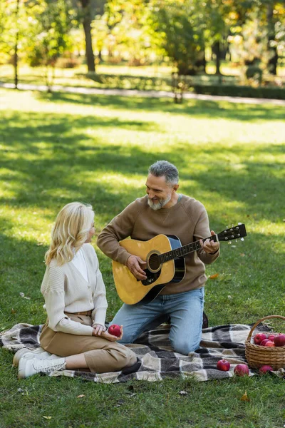 Счастливый мужчина средних лет с седой бородой играет на акустической гитаре рядом с блондинкой женой во время пикника в парке — стоковое фото
