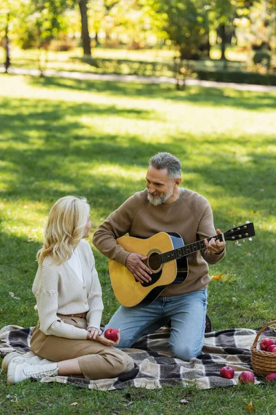 Allegro uomo di mezza età con barba grigia che suona la chitarra acustica vicino alla moglie bionda durante il picnic nel parco — Foto stock