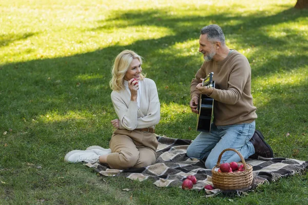 Happy middle aged man with grey beard playing acoustic guitar near carefree wife with apple during picnic in park — Stock Photo