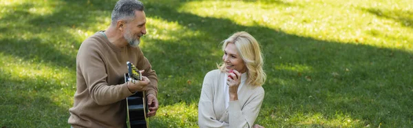 Happy middle aged man with grey beard playing acoustic guitar near carefree wife with apple in park, banner — Stock Photo