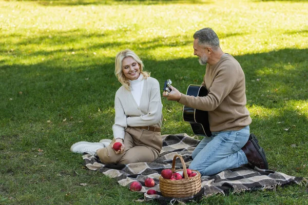 Feliz homem de meia-idade com barba cinza tocando guitarra acústica perto alegre esposa com maçã durante piquenique no parque — Fotografia de Stock
