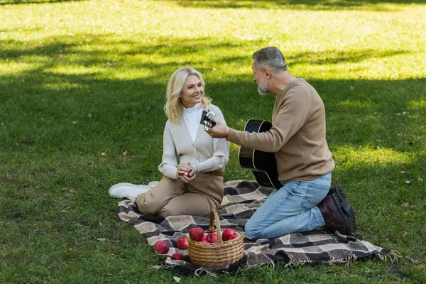 Homem de meia idade com barba cinza tocando guitarra acústica perto de esposa feliz com maçã durante piquenique no parque — Fotografia de Stock