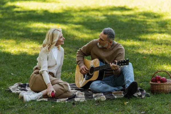 Мужчина средних лет с седой бородой играет на акустической гитаре рядом с очаровательной женой во время пикника в парке — стоковое фото