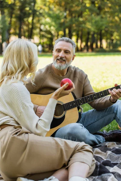 Радісний чоловік середнього віку з сірою бородою грає на акустичній гітарі біля блондинки з червоним яблуком під час пікніка в парку — стокове фото